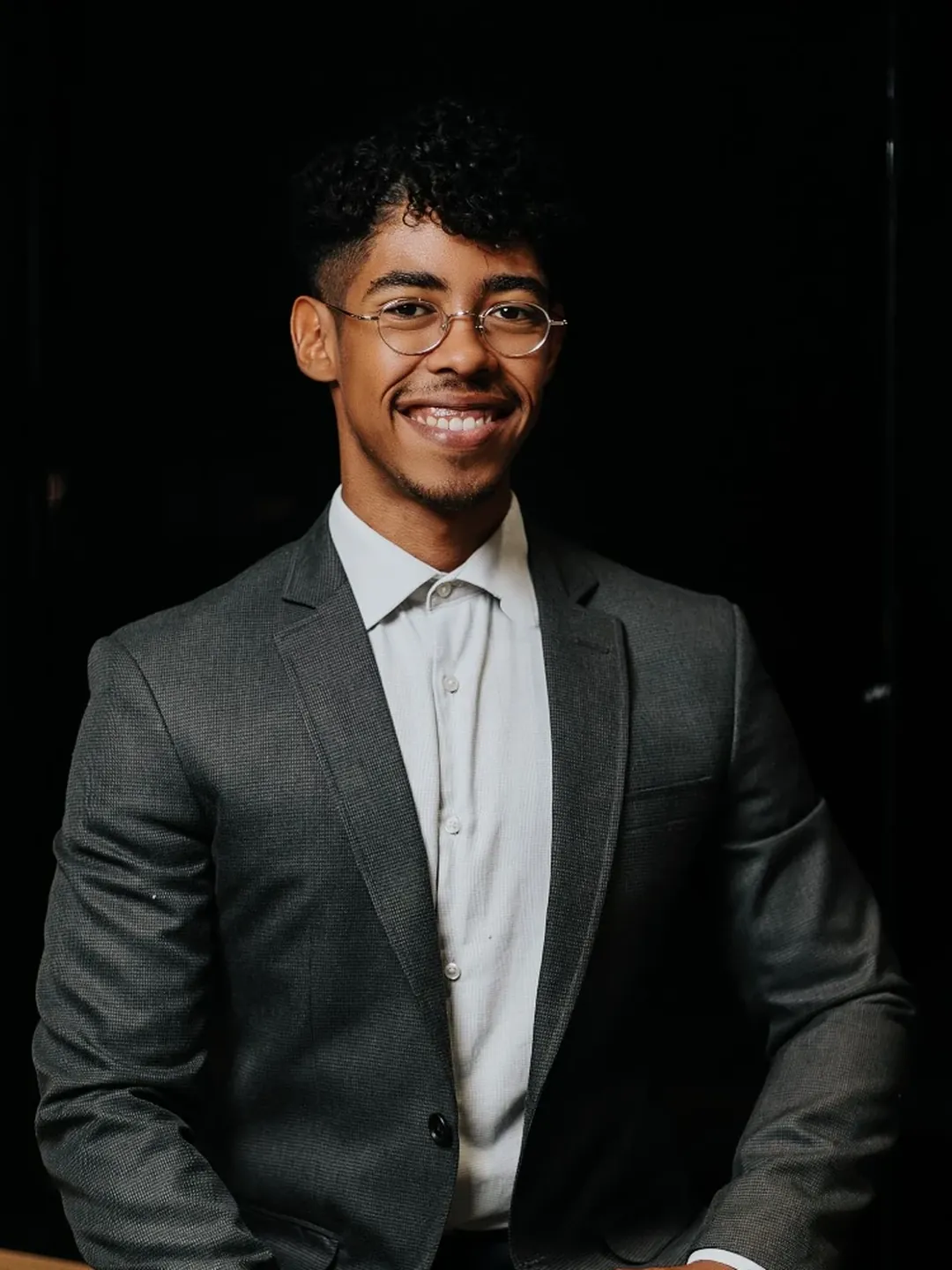 Efraim Vitaliano sorrindo, de óculos, blazer cinza e camisa clara, sentado em uma bancada de madeira
